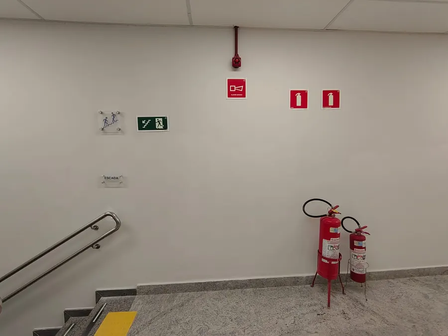 Escada interna de agência bancária com corrimão duplo, sinalização de rota de fuga, extintores e piso tátil de alerta.